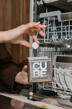 Hand holding a CUBE dishwasher cube above the reusable CUBE metal tin next to an open dishwasher