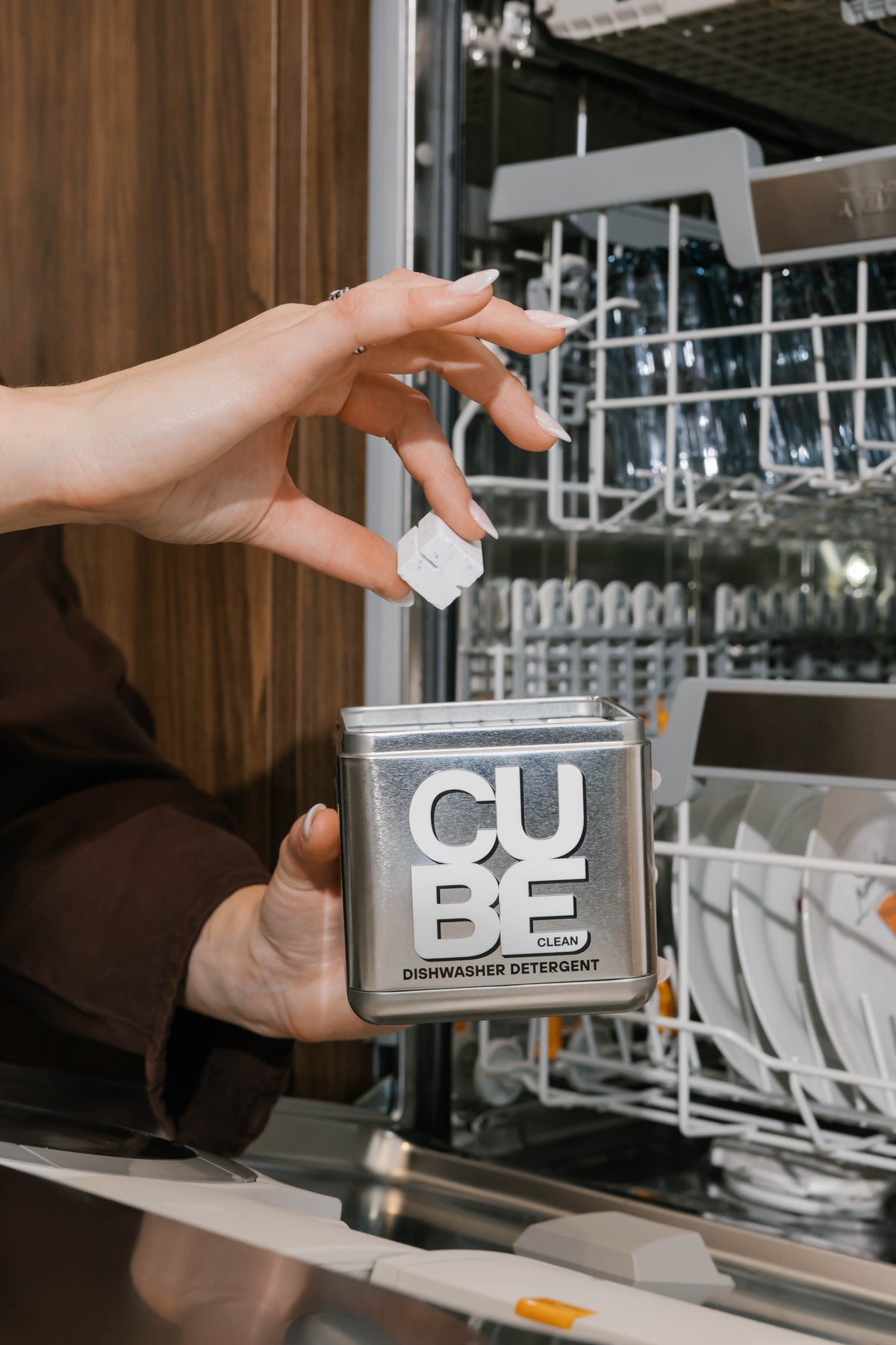 Hand holding a CUBE dishwasher cube above the reusable CUBE metal tin next to an open dishwasher